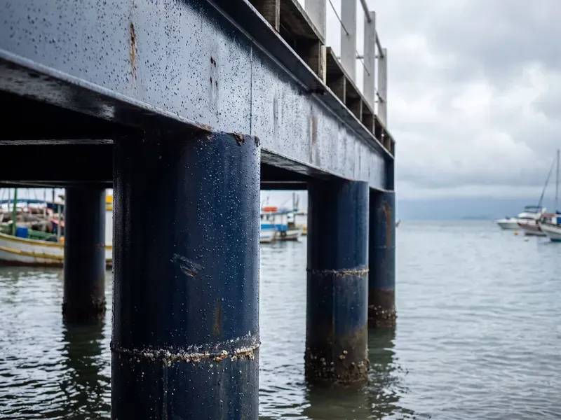 Estacas de aço de píer revestidas com primer epóxi escuro, linha de maré e cracas visíveis, mar e barcos ao fundo